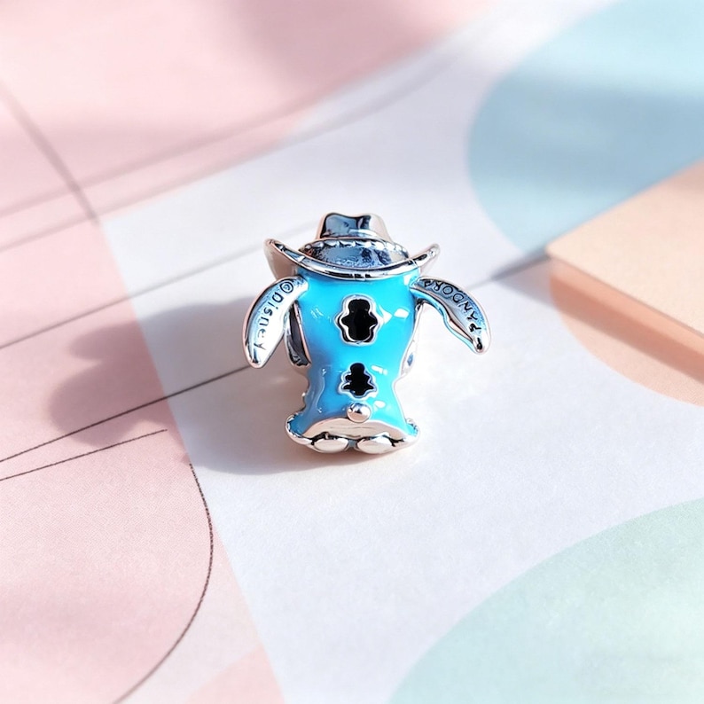 Puede incluir: Un charm de color azul claro con un personaje de dibujos animados con un sombrero de vaquero. El charm tiene detalles negros y la palabra "Disney" est&aacute; inscrita en un lado. El charm es de metal plateado, probablemente una joya.