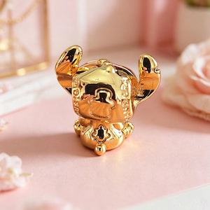 Peut inclure: Figurine dor&eacute;e d'un personnage de dessin anim&eacute; avec de grandes oreilles et un visage souriant. La figurine est tr&egrave;s r&eacute;fl&eacute;chissante, avec des d&eacute;tails complexes et une finition lisse. Le personnage est pos&eacute; avec les bras lev&eacute;s. La figurine est sur une surface rose.