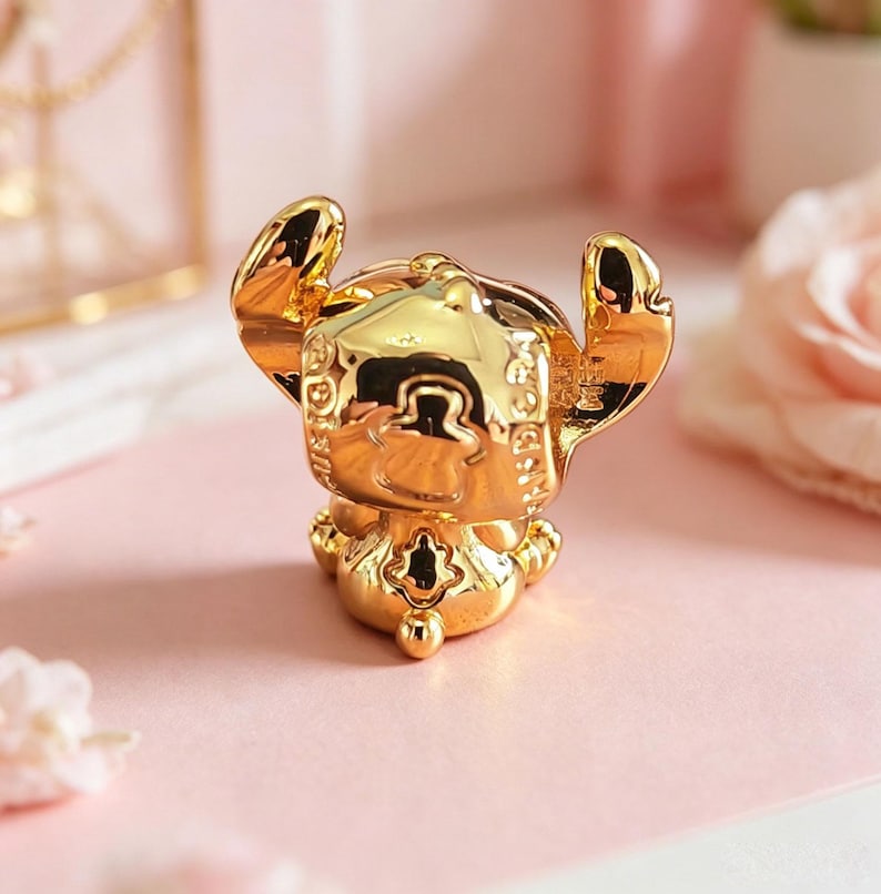 Peut inclure: Figurine dor&eacute;e d'un personnage de dessin anim&eacute; avec de grandes oreilles et un visage souriant. La figurine est tr&egrave;s r&eacute;fl&eacute;chissante, avec des d&eacute;tails complexes et une finition lisse. Le personnage est pos&eacute; avec les bras lev&eacute;s. La figurine est sur une surface rose.