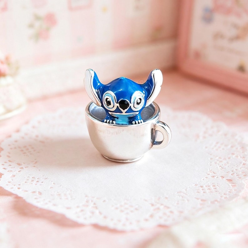Pode incluir: Uma figura azul de Stitch com detalhes brancos est&aacute; dentro de uma x&iacute;cara de ch&aacute; prateada. A figura tem olhos e orelhas grandes. A x&iacute;cara est&aacute; sobre uma toalha de renda branca, com um fundo rosa suave.