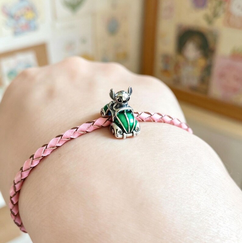 May include: A silver charm bracelet with a pink braided leather band. The charm is a small, detailed figure of a character in a green car. The charm has a green and silver color scheme.