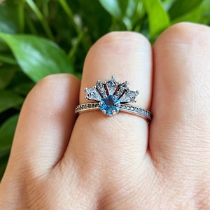Pode incluir: Um anel de prata com uma pedra preciosa azul redonda central, rodeada por uma disposição em forma de leque de pedras menores, claras e escuras. A banda é detalhada com pequenas pedras escuras. O anel é exibido num dedo.