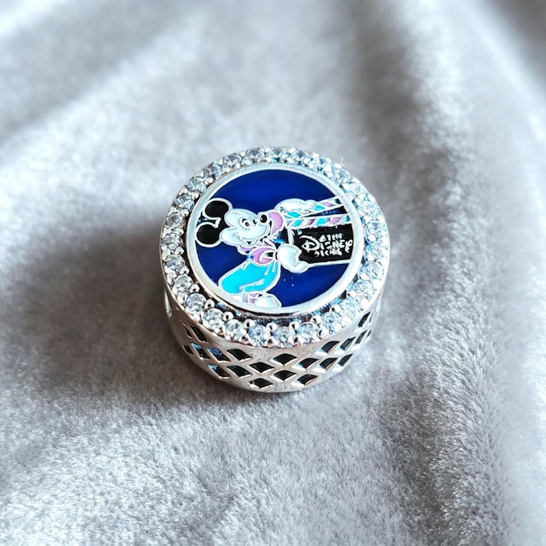 Pode incluir: Um berloque redondo prateado com um centro de esmalte azul apresentando Mickey Mouse segurando uma claquete de filme. O berloque &eacute; circundado por cristais transparentes e tem um design decorativo vazado nas laterais. O texto "Disney" &eacute; vis&iacute;vel.