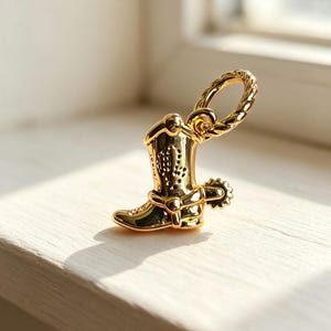 Könnte beinhalten: Ein polierter goldfarbener Metallanhänger in Form eines Cowboystiefels, der mit einem detaillierten Sporn an der Ferse und einer strukturierten, seilartigen runden Öse zur Befestigung versehen ist. Die Oberfläche ist glatt und reflektierend.