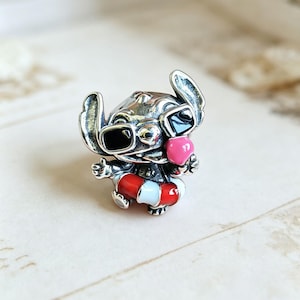 May include: A silver-toned metal charm featuring a character with large ears wearing black sunglasses and a pink nose. The character is positioned inside a red and white striped life preserver ring, with arms outstretched and a wide, open-mouthed expression.