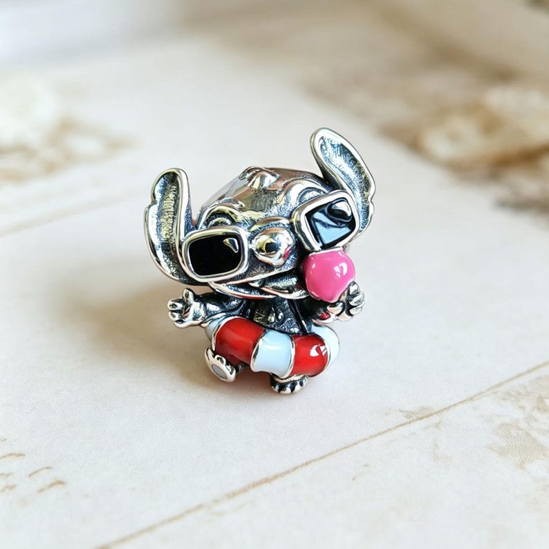 May include: A silver-toned metal charm featuring a character with large ears wearing black sunglasses and a pink nose. The character is positioned inside a red and white striped life preserver ring, with arms outstretched and a wide, open-mouthed expression.