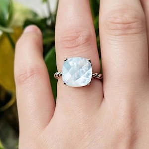 May include: A square-cut, iridescent gemstone ring with a twisted silver band. The gemstone has a light blue and white colour and is set in a four-prong setting. The ring is a piece of jewellery.