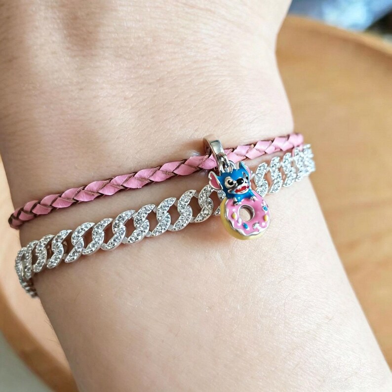 May include: Two bracelets are shown. The bottom bracelet is a silver chain with clear stones. The top bracelet is a pink braided leather. A charm with a blue character and a pink donut is attached to the top bracelet.