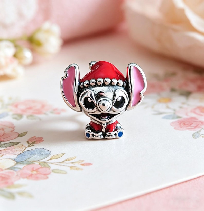 Op de afbeelding: Een zilveren bedel met een stripfiguur met een rode kerstmuts en outfit. De bedel heeft roze oren en blauwe accenten op de voeten. De achtergrond is zachtroze met bloemmotieven.