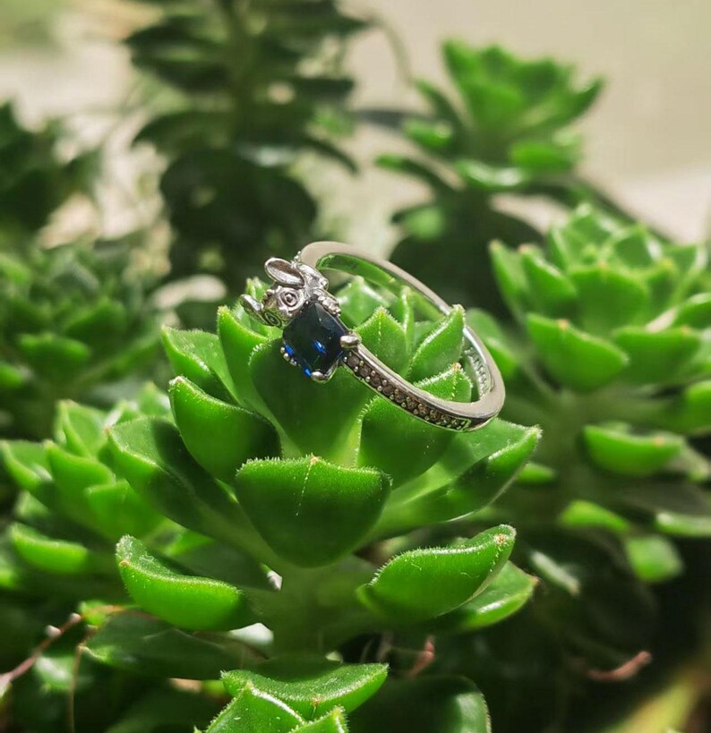 May include: A silver ring featuring a rabbit design with a dark blue, rectangular gemstone. The ring band is adorned with small, clear stones. The ring is set against a backdrop of green succulent plants.