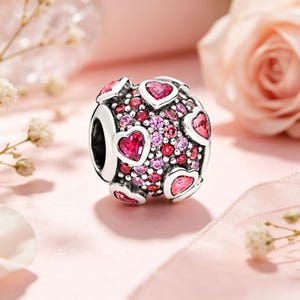 Puede incluir: Una cuenta de dije plateada con un tema de corazón. La cuenta está cubierta de pequeñas gemas rosas y rojas, con acentos más grandes en forma de corazón. El dije está diseñado para ser agregado a una pulsera o collar.