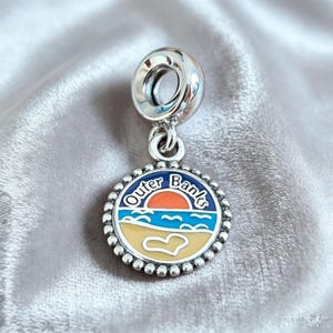 Könnte beinhalten: Ein silberfarbener Metallanhänger mit einem runden Anhänger mit Perlenrand. Die Mitte zeigt ein Emaille-Design einer Strandszene mit Sonnenuntergang, blauen Meereswellen, einem beigen Sandstrand mit weißem Herzumriss und dem Text Outer Banks.