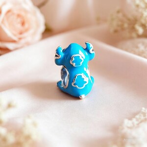 May include: A blue enamel charm in the shape of a cartoon character with silver accents. The charm features a light blue body with white cloud-shaped details and silver outlines. The charm is a collectible item.