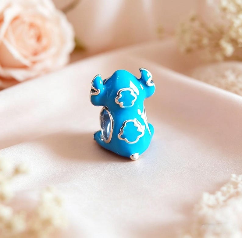 May include: A blue enamel charm in the shape of a cartoon character with silver accents. The charm features a light blue body with white cloud-shaped details and silver outlines. The charm is a collectible item.