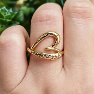Op de afbeelding: Een goudkleurige ring met een open, asymmetrisch hartvormig ontwerp. Het oppervlak van de band is versierd met talloze kleine, heldere, rondgeslepen stenen die in het gebogen metalen frame zijn gezet voor een sprankelende, textuurrijke uitstraling.