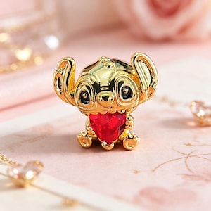 Peut inclure: Figurine dor&eacute;e d'un personnage de dessin anim&eacute; avec de grandes oreilles et une gemme rouge en forme de c&oelig;ur. La figurine a des yeux noirs et est assise. Le fond est rose p&acirc;le.