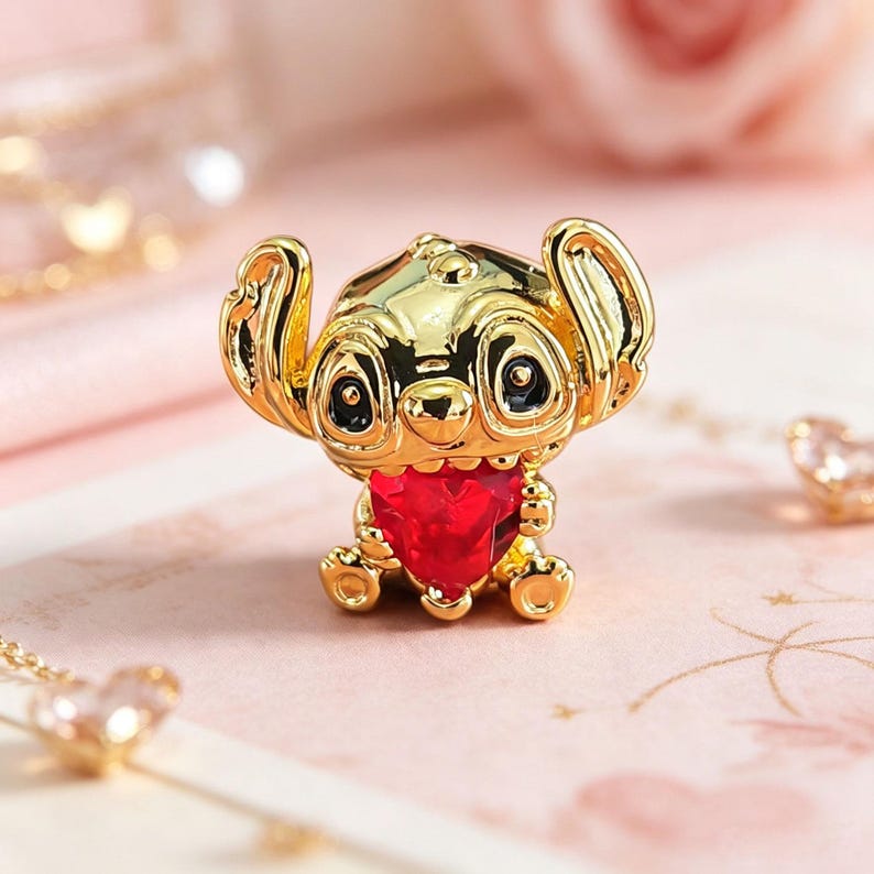 Peut inclure: Figurine dor&eacute;e d'un personnage de dessin anim&eacute; avec de grandes oreilles et une gemme rouge en forme de c&oelig;ur. La figurine a des yeux noirs et est assise. Le fond est rose p&acirc;le.