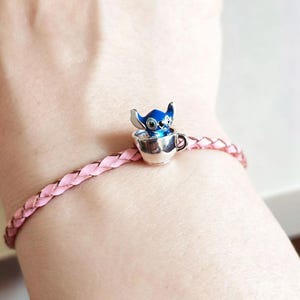 Pode incluir: Uma pulseira de couro tran&ccedil;ado rosa com um berloque prateado. O berloque apresenta um personagem Stitch azul e prateado em uma x&iacute;cara de ch&aacute; prateada. A pulseira &eacute; usada no pulso.