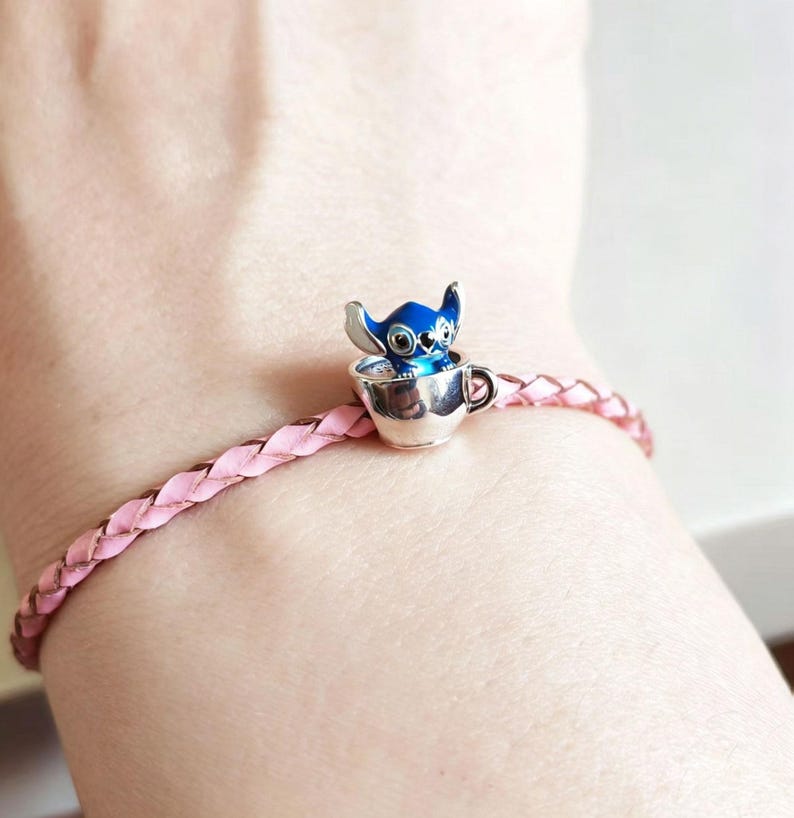 Pode incluir: Uma pulseira de couro tran&ccedil;ado rosa com um berloque prateado. O berloque apresenta um personagem Stitch azul e prateado em uma x&iacute;cara de ch&aacute; prateada. A pulseira &eacute; usada no pulso.
