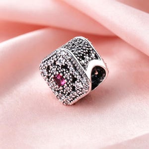 Pode incluir: Um berloque quadrado de prata com uma pedra preciosa rosa retangular central rodeada por numerosas pedras pequenas e claras cravadas em pavé. A superfície metálica tem um acabamento oxidado escuro nas áreas rebaixadas do design.
