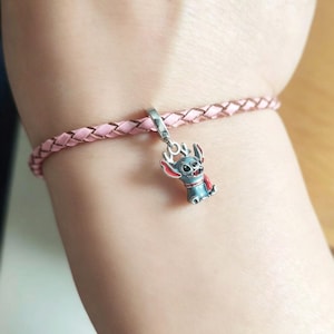 K&ouml;nnte beinhalten: Ein rosa geflochtenes Armband mit einem silbernen Anh&auml;nger, der eine blaue und rote Cartoonfigur zeigt. Der Anh&auml;nger ist eine stilisierte Figur mit gro&szlig;en Ohren und einem roten Umhang. Das Armband besteht aus hellrosa Material.