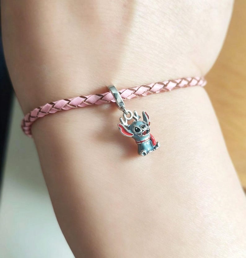 K&ouml;nnte beinhalten: Ein rosa geflochtenes Armband mit einem silbernen Anh&auml;nger, der eine blaue und rote Cartoonfigur zeigt. Der Anh&auml;nger ist eine stilisierte Figur mit gro&szlig;en Ohren und einem roten Umhang. Das Armband besteht aus hellrosa Material.