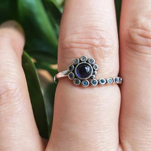 Pode incluir: Um anel de prata com uma pedra central circular azul-escura rodeada por uma espiral de pedras mais pequenas de cor azul-clara. A banda de metal tem um acabamento polido e segue um design em espiral que se estreita ao longo do dedo.