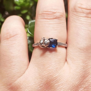 May include: Silver ring featuring a square, deep blue gemstone. The ring has a detailed design on the band and a small, decorative element on one side. The band is adorned with small, clear stones.