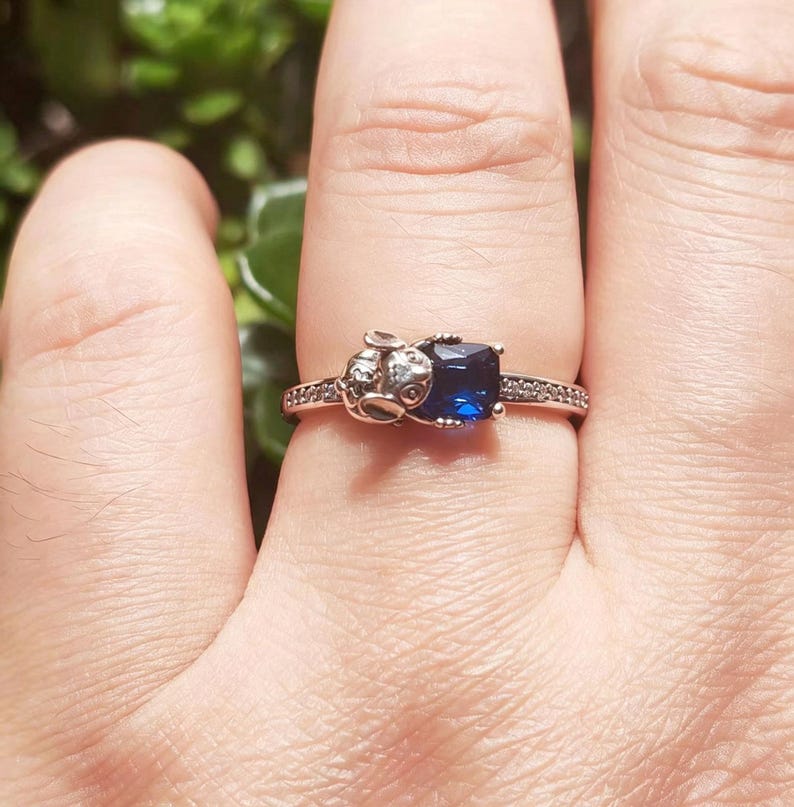 May include: Silver ring featuring a square, deep blue gemstone. The ring has a detailed design on the band and a small, decorative element on one side. The band is adorned with small, clear stones.