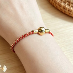 Puede incluir: Una pulsera que presenta un cordón rojo fino entrelazado con una cadena metálica de tono dorado. El diseño incluye una cuenta esférica central de tono dorado pulido flanqueada por dos pequeños conectores cilíndricos dorados.