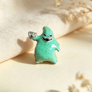Può includere: Una spilla in smalto verde menta raffigurante un personaggio con un ampio sorriso scuro e frastagliato e occhi stretti. La figura ha una finitura glitterata e tiene in mano due piccoli dadi quadrati color argento, delineati da un bordo in metallo.
