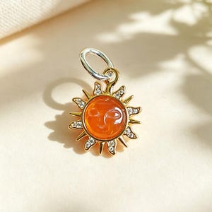 Peut inclure: Un pendentif soleil doré avec un cabochon orange central présentant un visage en relief subtil. Les rayons du soleil sont ornés de petites pierres claires et scintillantes. Un anneau argenté est fixé au sommet de la breloque en forme de soleil.