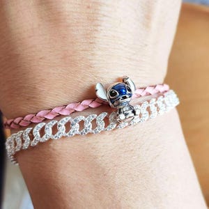 May include: A silver-colored charm bracelet with a cartoon character charm and a pink braided bracelet. The charm is a blue and white character. The bracelets are worn on a wrist.