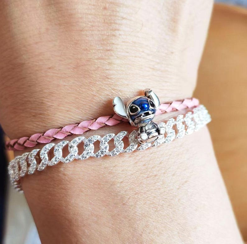 May include: A silver-colored charm bracelet with a cartoon character charm and a pink braided bracelet. The charm is a blue and white character. The bracelets are worn on a wrist.