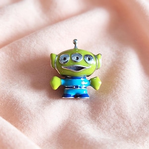 Peut inclure: Petite figurine d'extraterrestre vert à trois yeux, avec des détails argentés et une tenue bleue et verte. La figurine a une bouche souriante et se tient debout, les bras tendus. Le jouet est sur une surface rose.