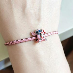 Pu&ograve; includere: Bracciale in pelle intrecciata rosa chiaro con un charm di Stitch rosa con occhi blu e un bastoncino di zucchero. Il charm presenta dettagli rossi, bianchi e argentati. Il bracciale &egrave; di colore rosa chiaro.