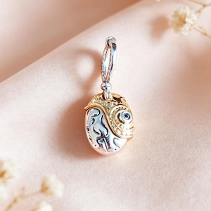 May include: A two-toned charm pendant featuring a polished silver-coloured oval base with embossed abstract patterns and a textured gold-coloured overlay on the right side, accented with a small embedded dark stone and a silver-coloured jump ring.