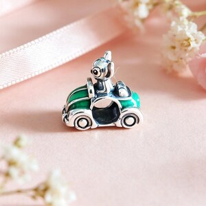 May include: A silver charm featuring a small dog driving a green convertible car. The car has silver wheels and trim. The dog has large eyes and is wearing a helmet. The charm is set against a soft pink background with flowers.