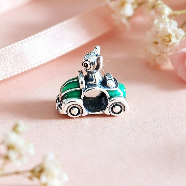 May include: A silver charm featuring a small dog driving a green convertible car. The car has silver wheels and trim. The dog has large eyes and is wearing a helmet. The charm is set against a soft pink background with flowers.