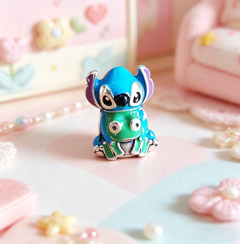 May include: A blue and green enamel charm featuring a cartoon character. The charm has a blue head with purple ears and a green body. The charm is sitting on a pink and blue surface.