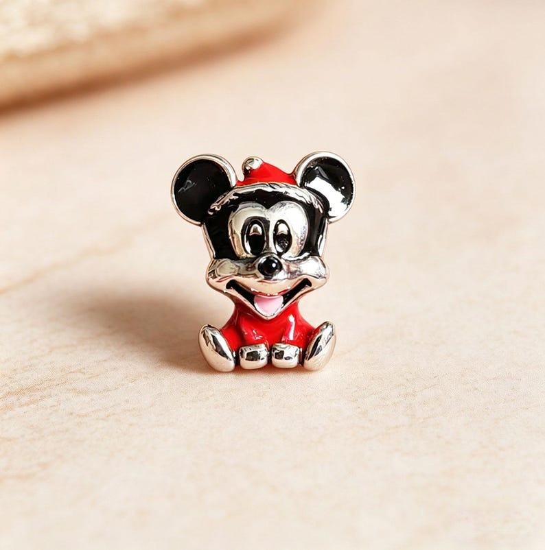 K&ouml;nnte beinhalten: Ein silberner Charm-Anh&auml;nger mit einer sitzenden Mickey Mouse-Figur. Die Figur tr&auml;gt ein rotes Outfit und einen Weihnachtsm&uuml;tze. Der Charm hat schwarze Ohren und ein l&auml;chelndes Gesicht mit rosa Zunge.