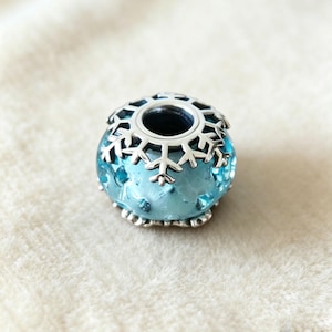 Peut inclure: Une perle en verre bleu avec un motif de flocon de neige. La perle a un dessus et une base en métal argenté. Le verre a un aspect texturé avec de petits points en relief. Le motif de flocon de neige est répété sur le dessus de la perle.