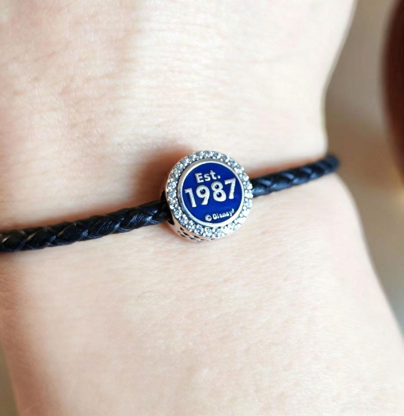 Pode incluir: Uma pulseira de couro tran&ccedil;ado preta com um berloque redondo. O berloque tem um centro de esmalte azul com o texto "Est. 1987" e &eacute; rodeado por um anel de cristais transparentes. A pulseira &eacute; usada em um pulso.