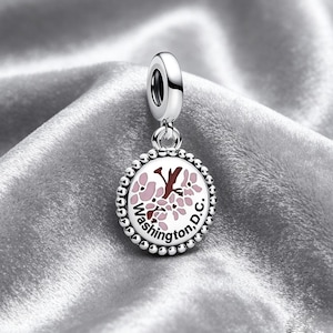 May include: A silver charm with a round pendant. The pendant displays a pink and brown floral design and the text "Washington, D.C.". It has a beaded border and a loop for attaching to a bracelet or necklace.