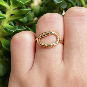 Può includere: Un anello color oro caratterizzato da un elemento centrale ovale dalla forma organica e irregolare. La superficie dell'ovale è impreziosita da diverse piccole pietre trasparenti a taglio rotondo incastonate a intervalli sulla montatura.