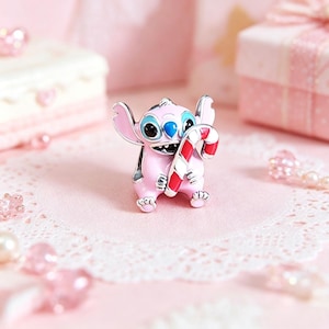 Pu&ograve; includere: Un charm rosa e argento raffigurante un personaggio dei cartoni animati con un bastoncino di zucchero rosso e bianco. Il charm ha occhi neri con dettagli blu, su uno sfondo rosa con elementi decorativi.