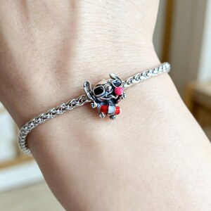May include: A silver-toned chain bracelet featuring a small, detailed charm of a blue creature with large ears, wearing black sunglasses, a bright pink nose, and a red and white life preserver ring around its midsection, secured by a metal clasp.