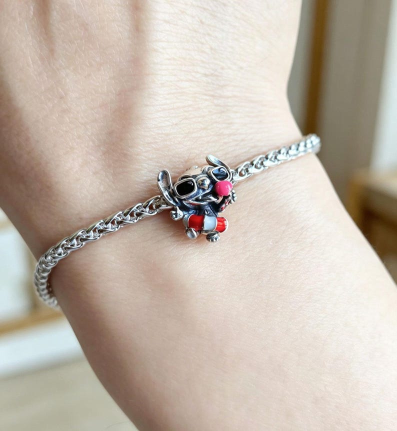 May include: A silver-toned chain bracelet featuring a small, detailed charm of a blue creature with large ears, wearing black sunglasses, a bright pink nose, and a red and white life preserver ring around its midsection, secured by a metal clasp.