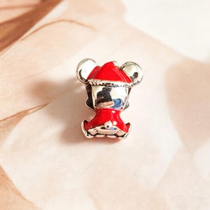 K&ouml;nnte beinhalten: Ein silberner und roter Charm-Anh&auml;nger mit einer Cartoon-Figur, die einen Weihnachtsmannhut und -outfit tr&auml;gt. Der Charm hat gro&szlig;e silberne Ohren und ein rotes Outfit mit wei&szlig;em Besatz. Der Charm ist wahrscheinlich aus Metall und Emaille.