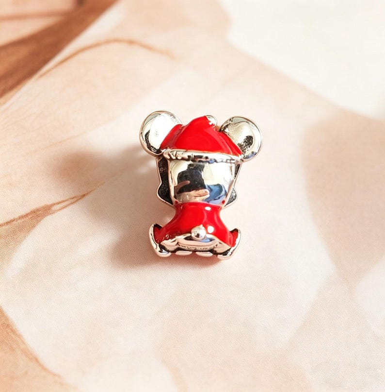 K&ouml;nnte beinhalten: Ein silberner und roter Charm-Anh&auml;nger mit einer Cartoon-Figur, die einen Weihnachtsmannhut und -outfit tr&auml;gt. Der Charm hat gro&szlig;e silberne Ohren und ein rotes Outfit mit wei&szlig;em Besatz. Der Charm ist wahrscheinlich aus Metall und Emaille.